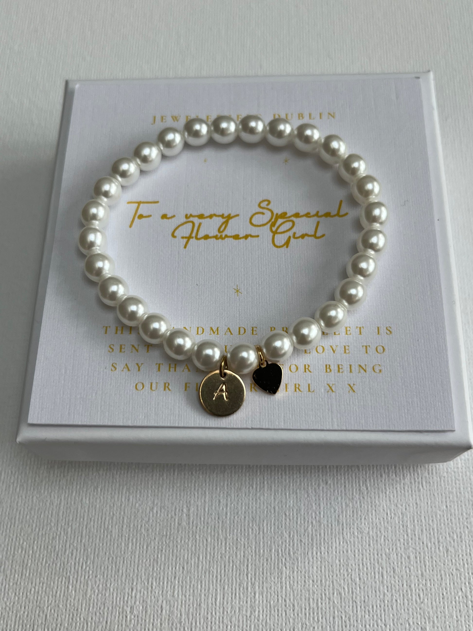 A pearl beaded flower girl bracelet with a gold initial 'A' charm and a small black heart charm, displayed on a white gift box with gold lettering.